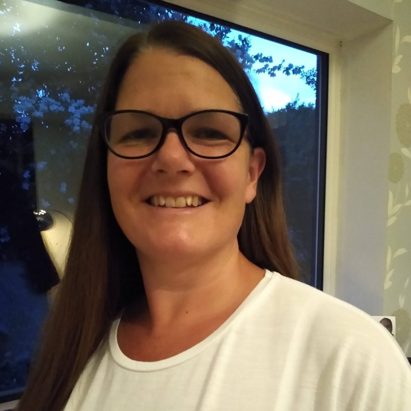Julie Smiling woman with long brown hair, wearing glasses, in a well-lit room.